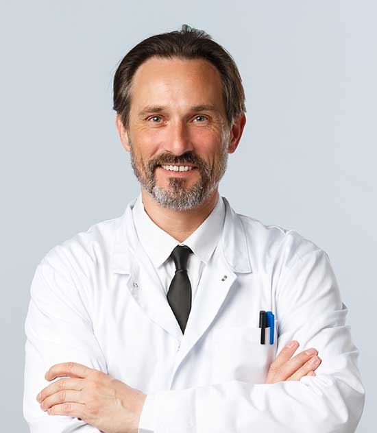 Smiling male doctor in a white coat with arms crossed, wearing a black tie and pens in his pocket.